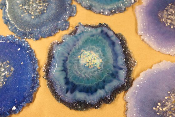 A close-up overhead view of several finished resin geode coasters in various shades of blue, turquoise, and purple, featuring jagged, glittery edges and textured, crystalline centers. The coasters are resting on a light brown surface.